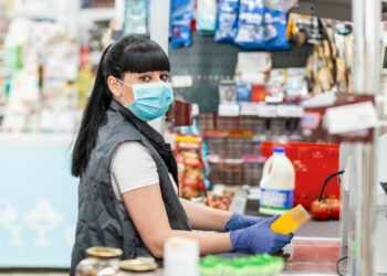A young woman in a medical mask and gloves, working at the checkout in a supermarket. Concept of coronovirus, protection from infection and industrial crisis.